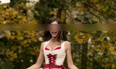 SP Escort Agency Munich, Jessica in a beige-red Dirndl with highheels. Shes swinging her skirt whilst smiling, in a garden.